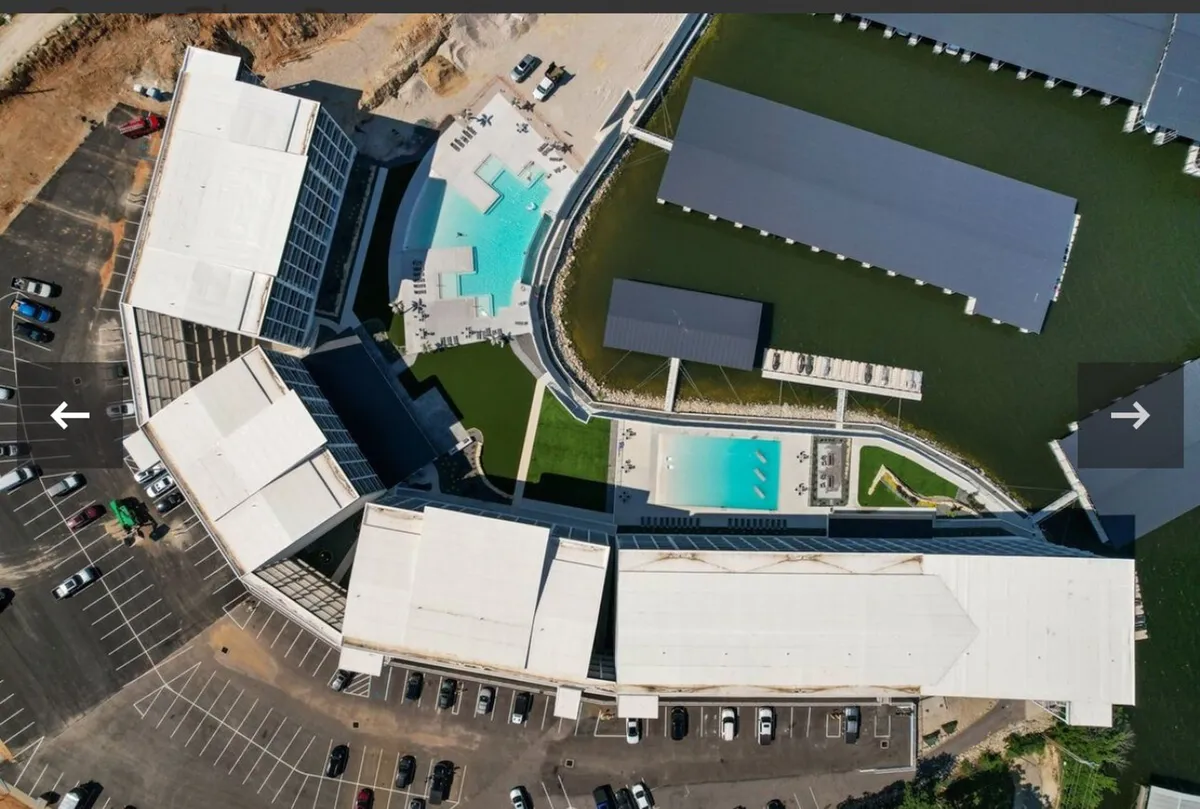 An expansive exterior view featuring the resort-style swimming pools, a dock area stretching into the lake, modern buildings in the background, and well-maintained green spaces around the pool. The scene is lively, with people swimming, sunbathing, and gathering near the dock, evoking a relaxed yet vibrant resort ambiance.