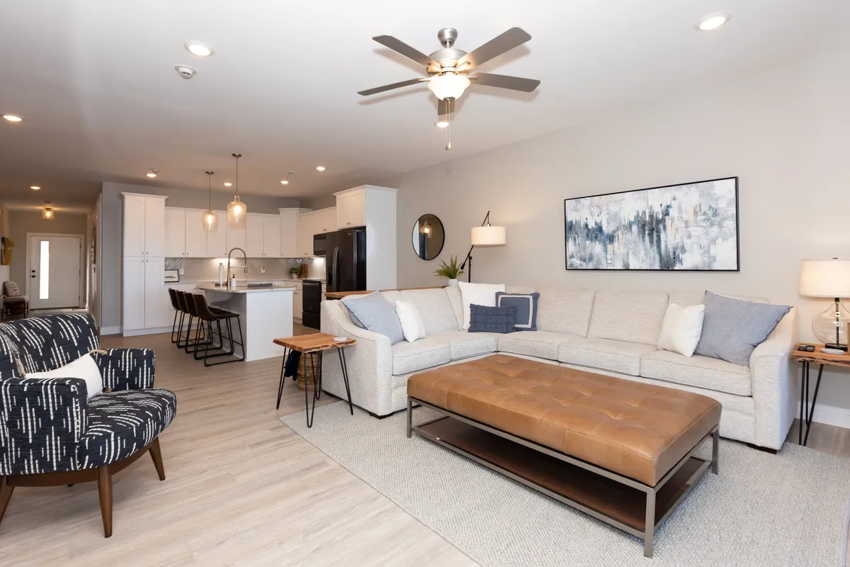 A welcoming living room with a plush sofa, modern coffee table, artwork, ceiling fan, and sightlines to the nearby kitchen area, all bathed in soft natural light. The space is arranged for both relaxation and togetherness.