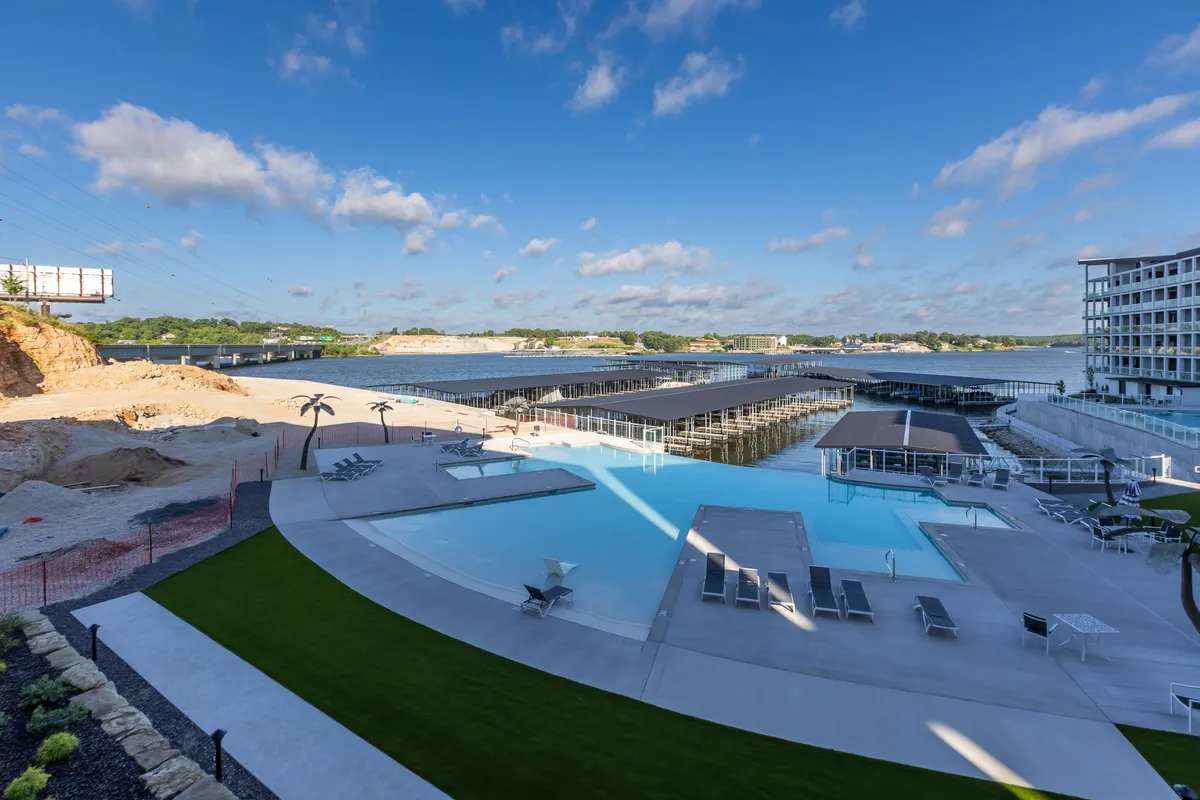 A vibrant exterior scene showcases a sparkling swimming pool surrounded by sun loungers and lush landscaping, with a marina and the expansive lake visible just beyond. The image conveys easy access to both the pool and the water, with plenty of space for relaxation, sunbathing, and water activities.