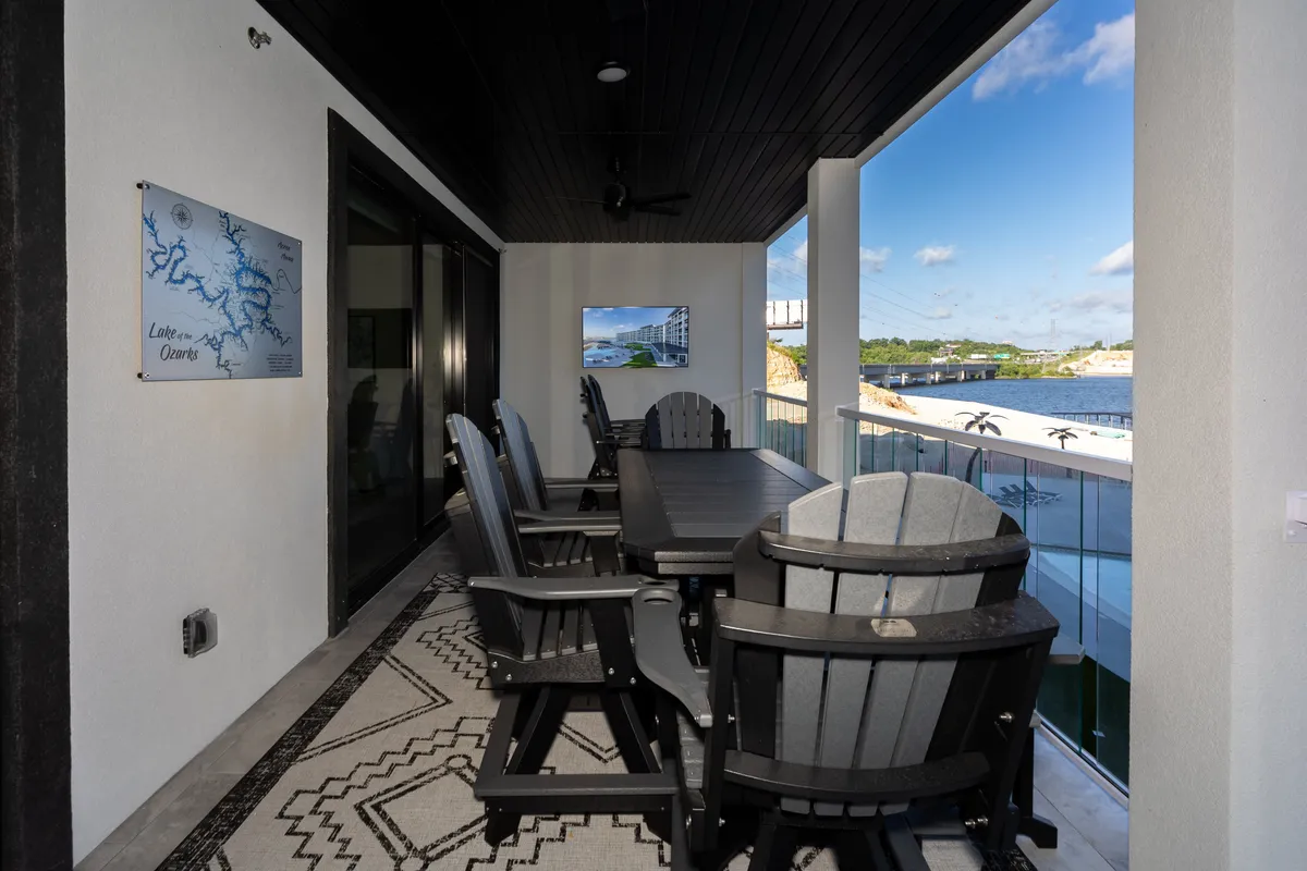 A private balcony set for outdoor dining, complete with Adirondack chairs, a table, a wall-mounted TV, and sweeping water views. The setup invites relaxing with coffee or cocktails while enjoying the sunset.
