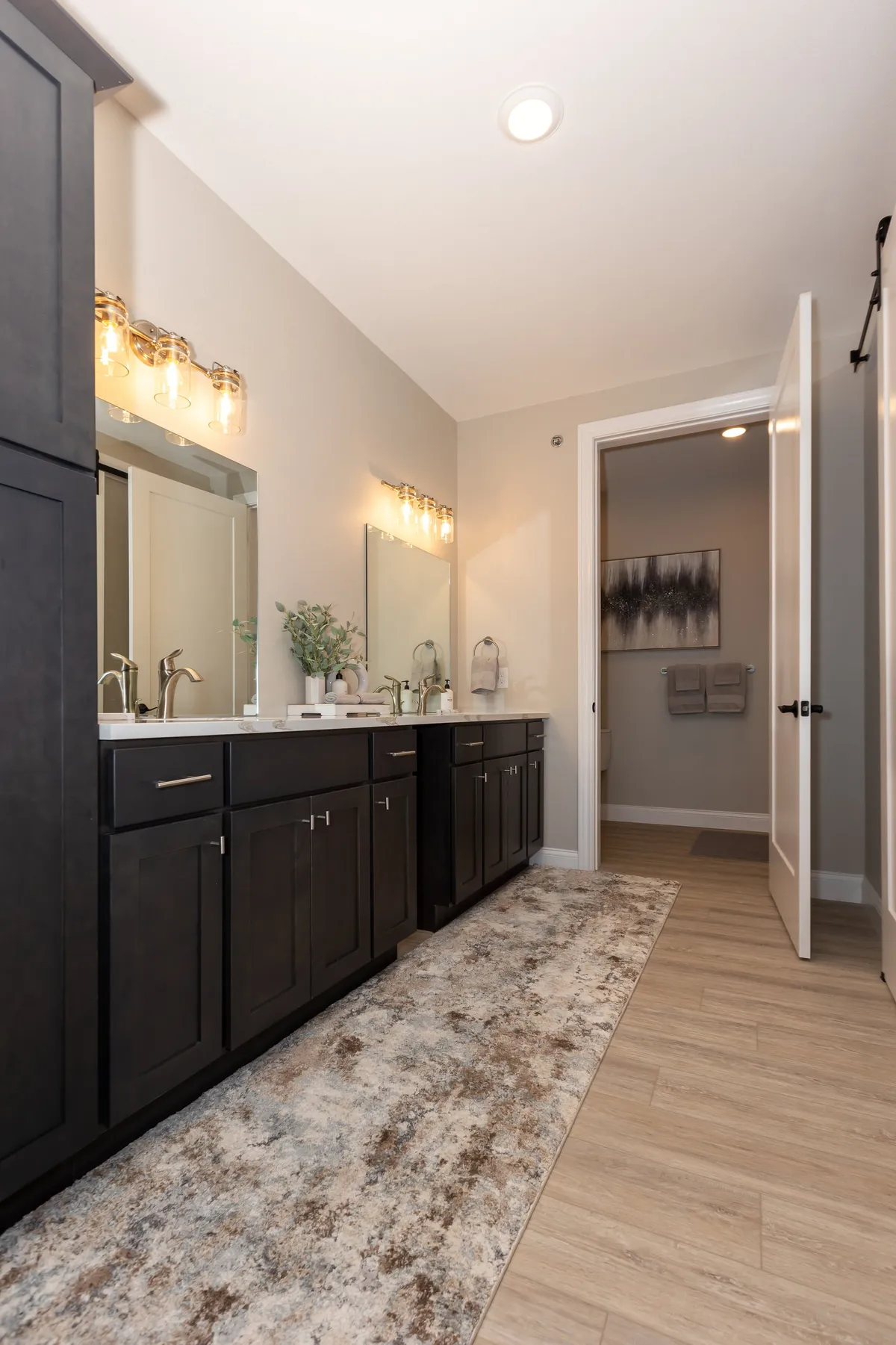 A modern bathroom with a double vanity, large mirrors, decorative lighting, and a soft rug, bathed in gentle morning light. The design is serene and inviting, reflecting a spa-like ambiance.
