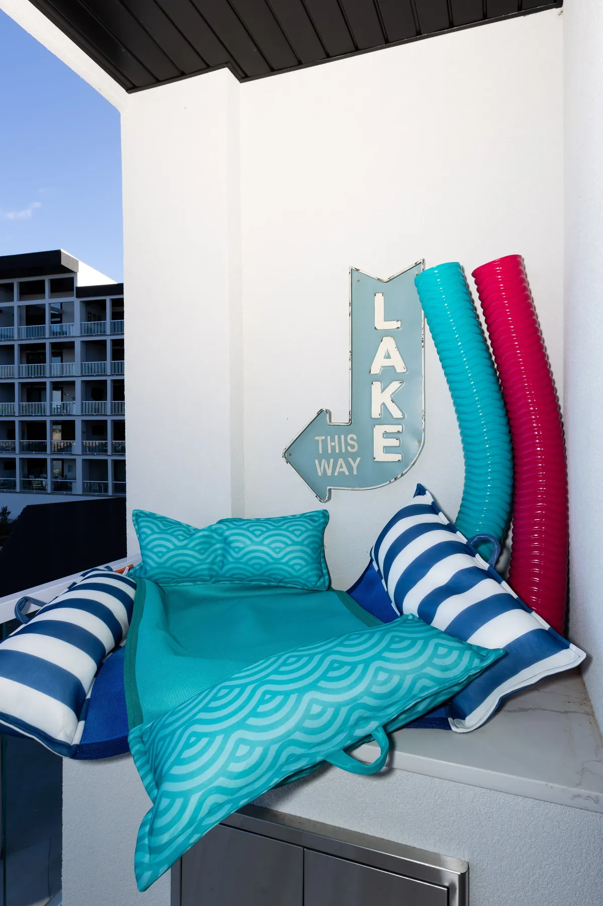 A cheerful lakeside setting with a colorful 'Lake' sign, striped outdoor cushions, and a collection of brightly colored pool noodles and floaties. The backdrop includes the tranquil lake, suggesting easy access for aquatic fun and relaxation.