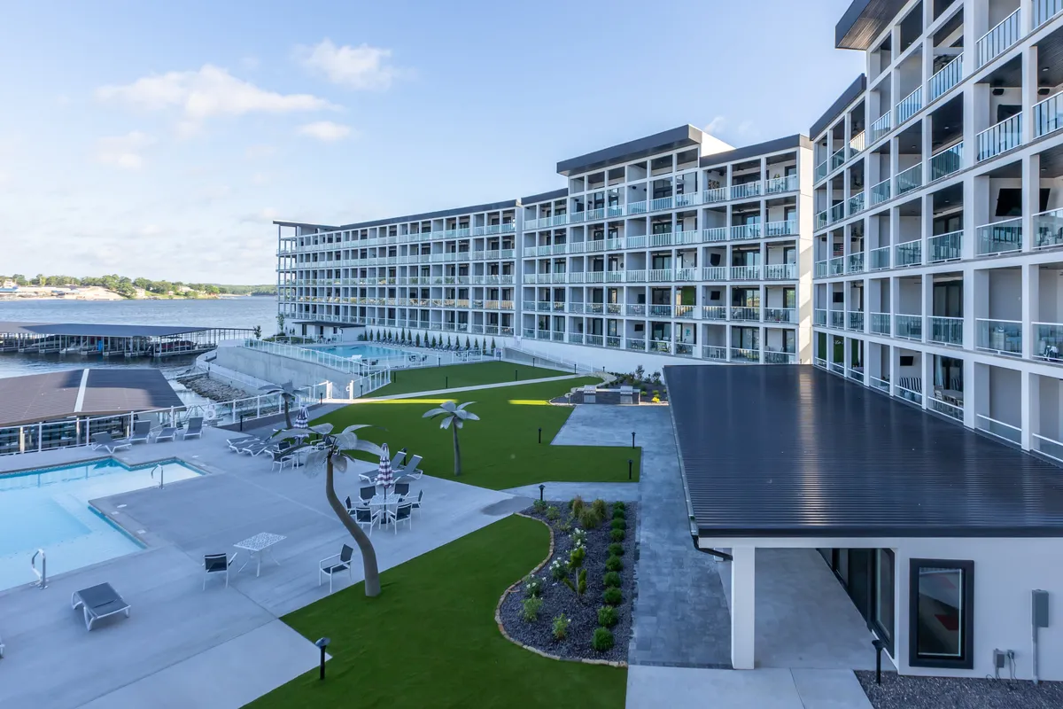 The main building of the property sits beside a large, tranquil pool area, surrounded by landscaped gardens and a waterfront. The scene conveys a resort-style atmosphere with plenty of outdoor space for relaxation and family enjoyment.