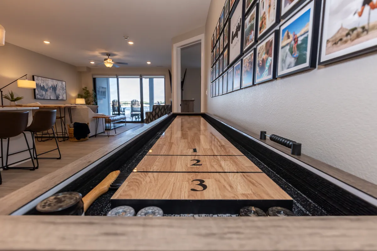 A view of the shuffleboard table positioned near a gallery wall of family photos, with the dining area visible in the background. The setup is casual yet stylish, emphasizing opportunities for friendly competition and shared meals.