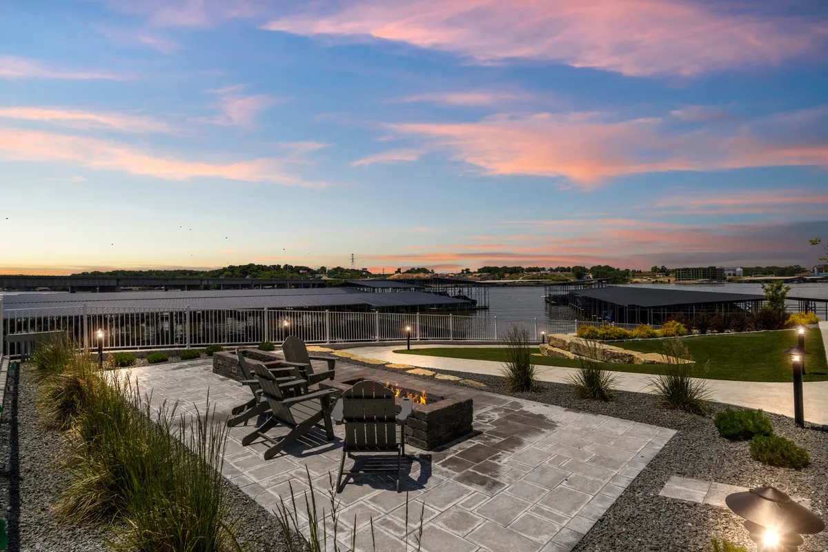 An inviting outdoor fire pit surrounded by Adirondack-style chairs, all set on a spacious patio overlooking the tranquil lake. The sun is setting in the background, casting a warm glow across the water, and the setup evokes relaxation and the pleasures of lakeside evenings.