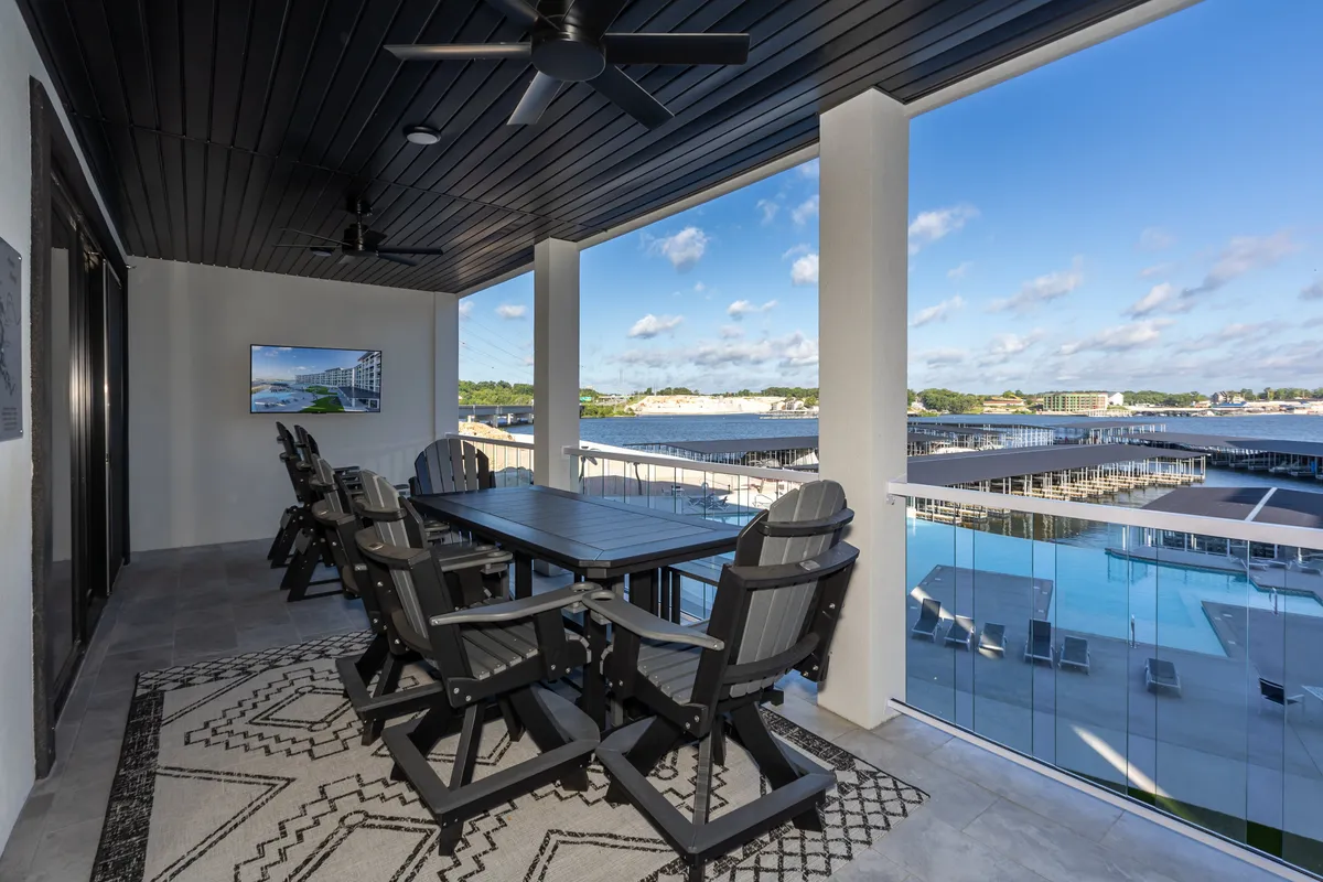 The image showcases an inviting outdoor dining area on a spacious deck, overlooking the tranquil waters of Osage Beach. A stylish dining table set atop a decorative rug sits under a ceiling fan for comfort, offering breathtaking lake views. The setup is perfect for sunset meals, relaxed gatherings, and watching boats drift by in the golden evening light.