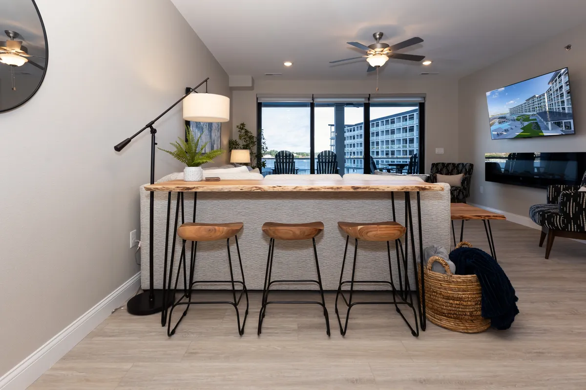 A kitchen island with wooden top and bar stools, flanked by large windows offering expansive views of the lake and resort amenities outside. Decorative plants and natural light enhance the bright, communal atmosphere.